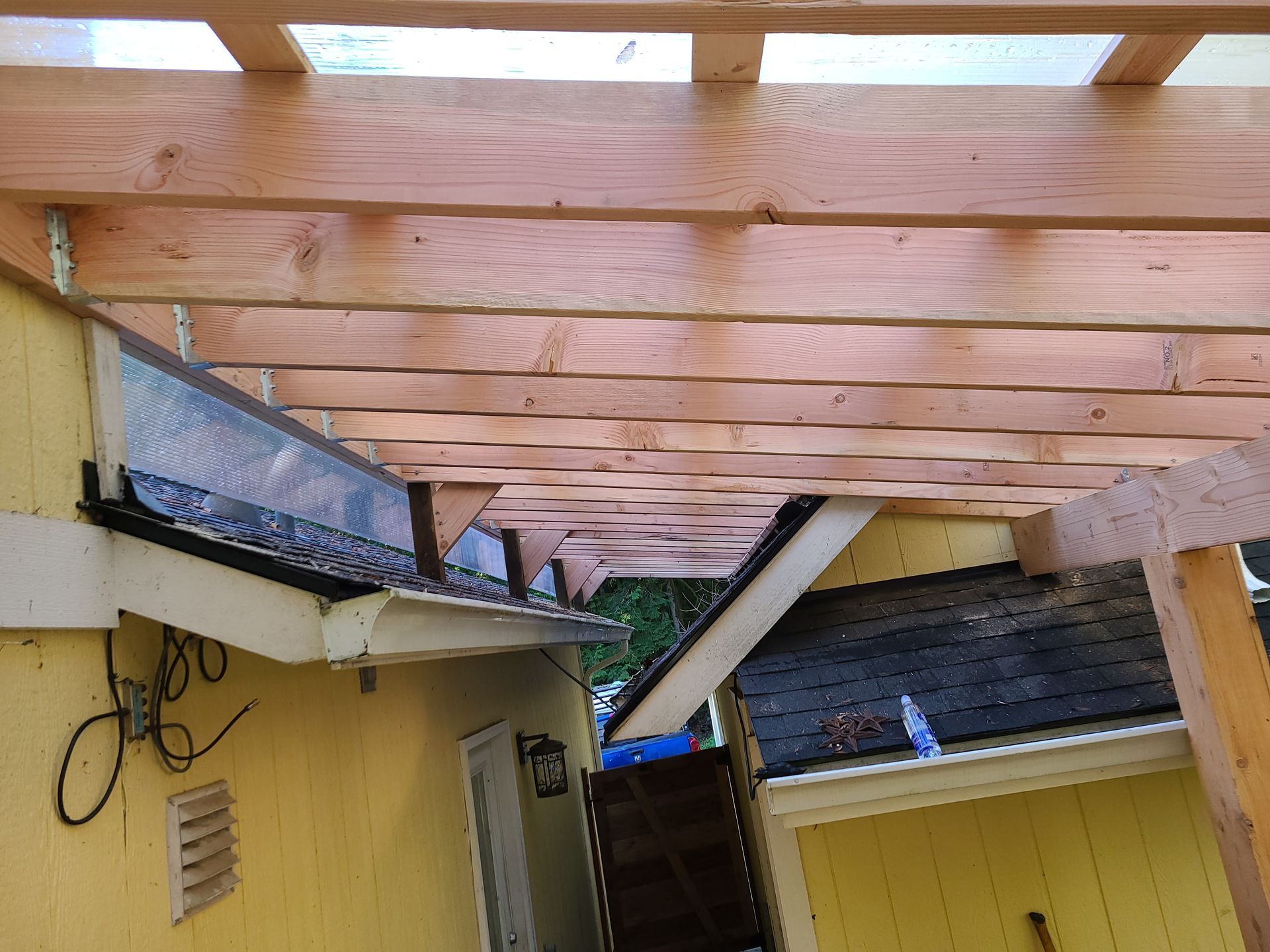 Wooden pergola attached to a yellow building, with a gutter visible.