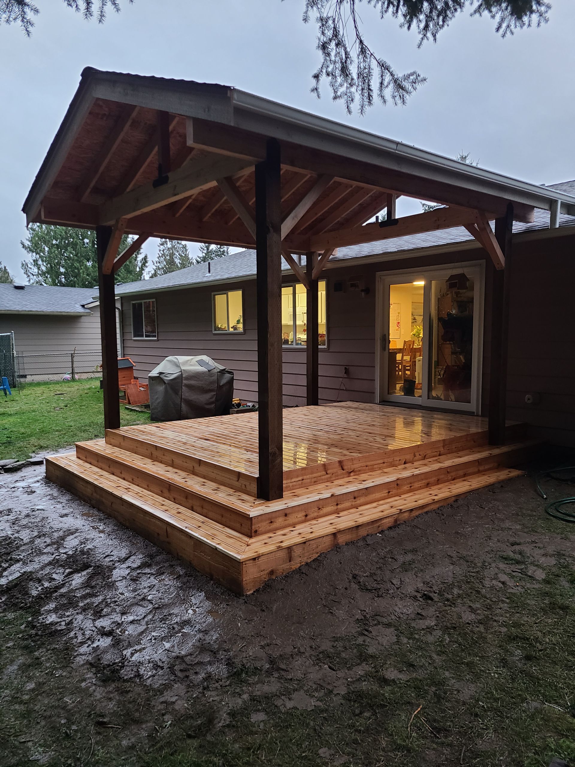 Wooden deck with steps and a covered roof attached to a house. Exterior setting with wet ground.