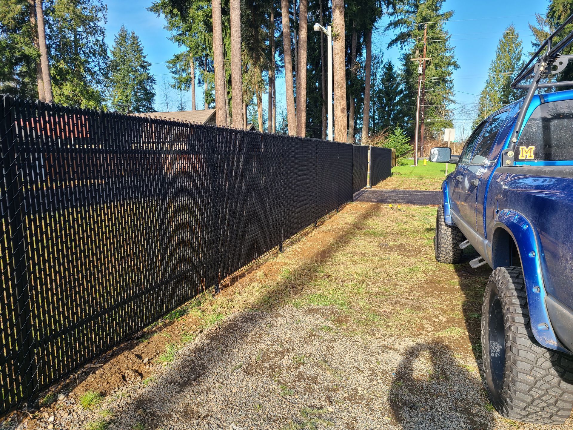 Black decorative chain-link fence along gravel with a blue truck parked on the right. Trees in the background.