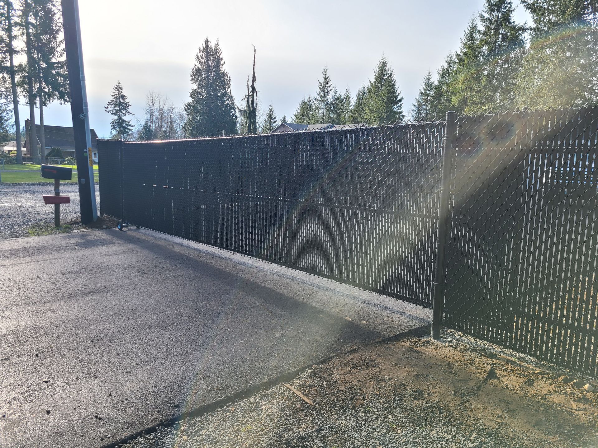 Black chain-link fence with privacy slats, blocking driveway entrance. Trees in background, bright sunlight.