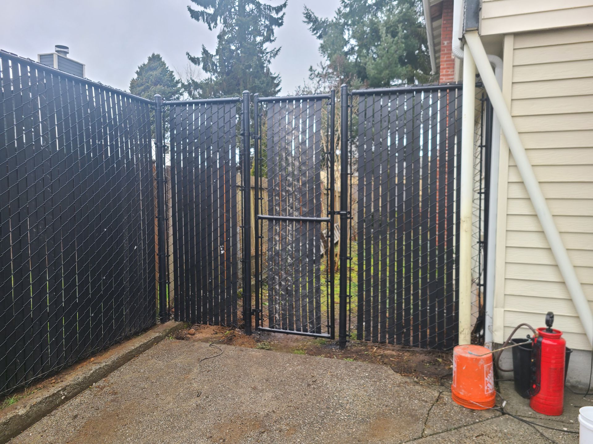 Black fence with gate on a concrete patio. Orange bucket and fire extinguisher nearby.