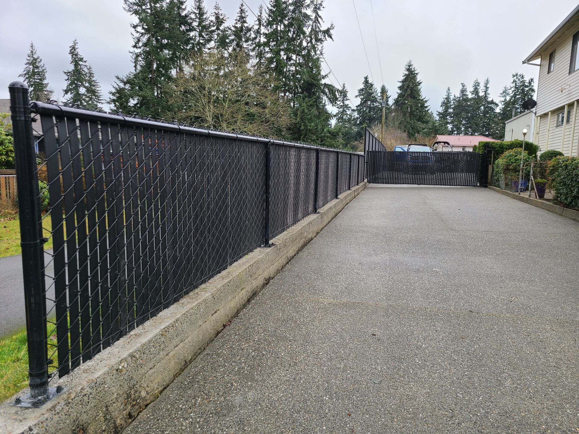 Black chain-link fence on a concrete base, separating a gravel surface from a residential area with trees.
