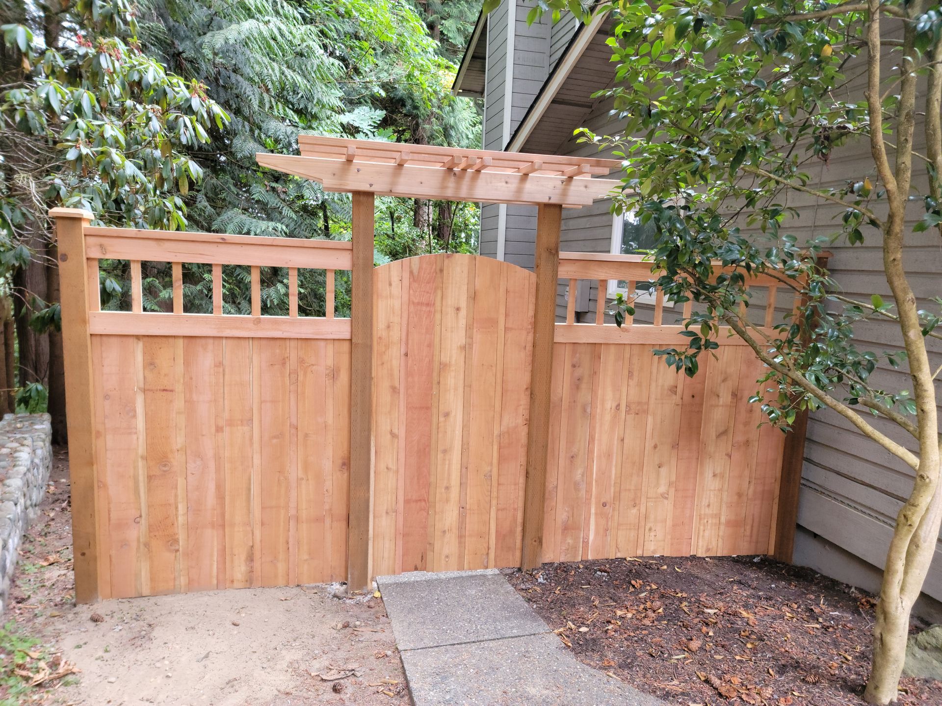 Wooden gate in a cedar fence with a pergola-like top, pathway, and surrounding greenery.