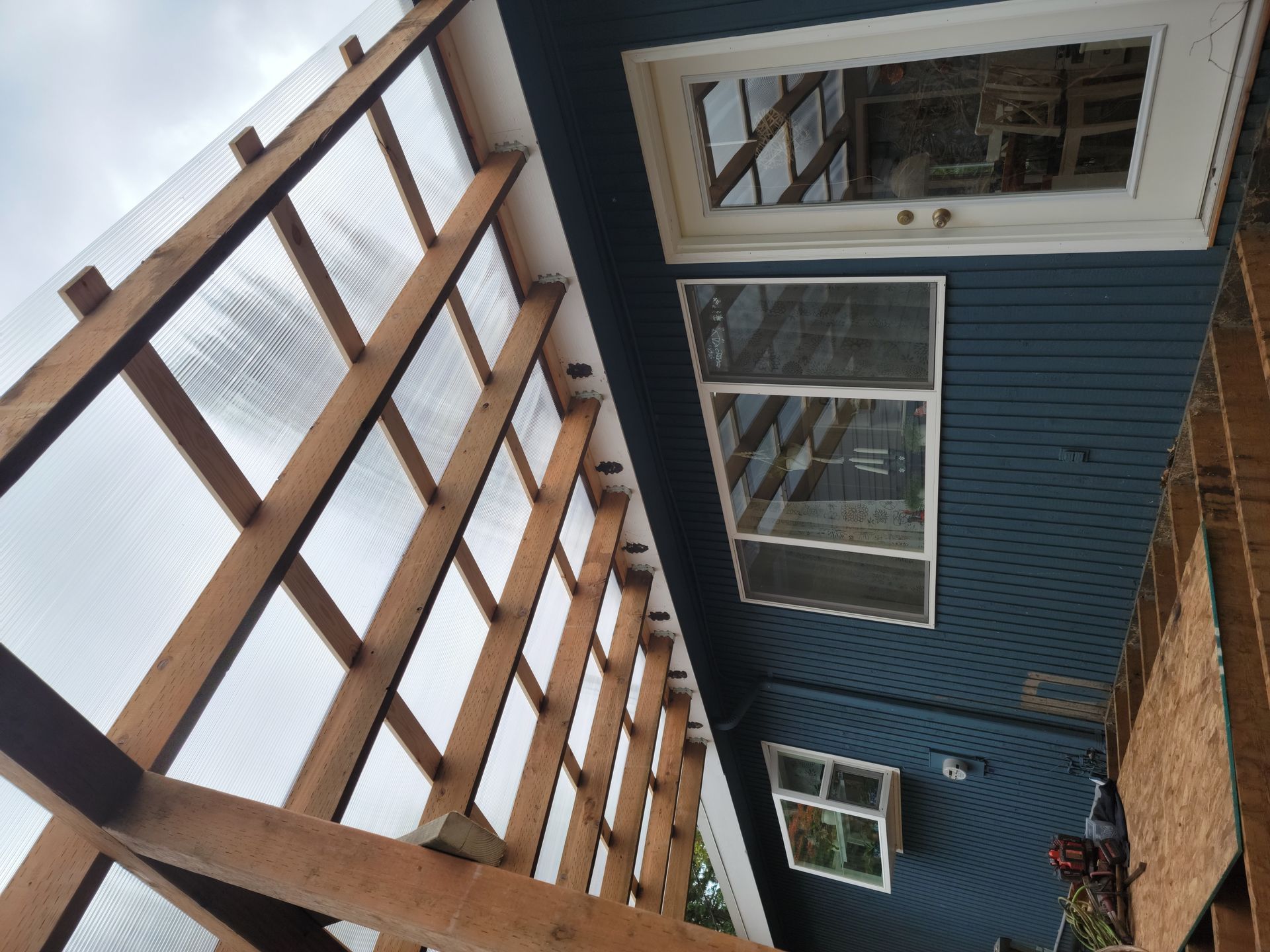 Wooden structure with clear roof panels attached to a blue house with windows.