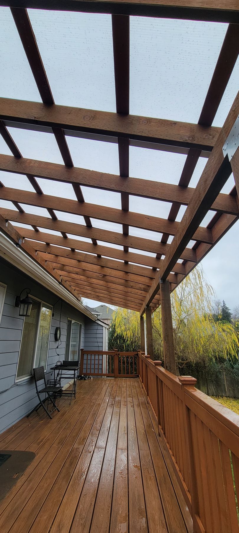 Wooden deck and pergola on a rainy day. Light-colored wooden railing and house siding.