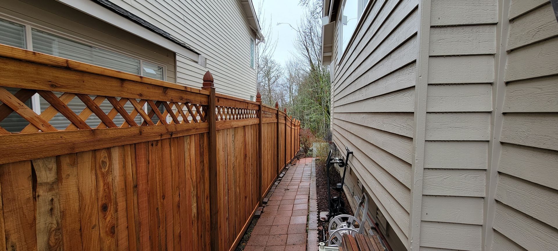 A narrow brick pathway between a wooden fence and a house with beige siding. Overcast sky.