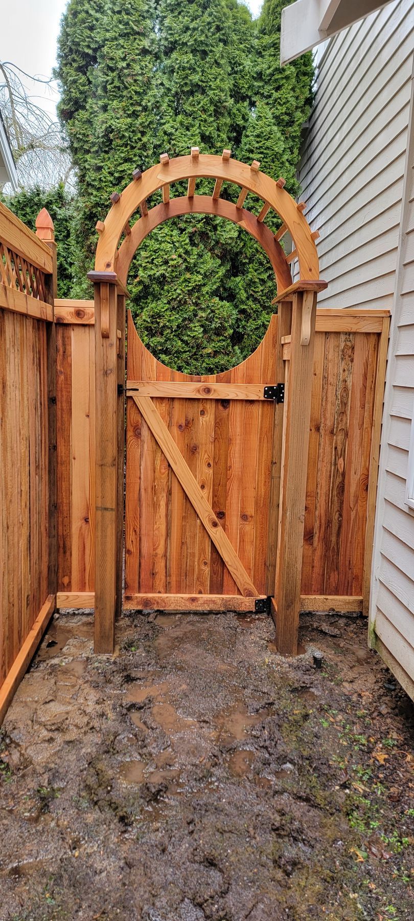 Wooden fence with arched gate. Gate is open, showing greenery behind. Brown soil in foreground.
