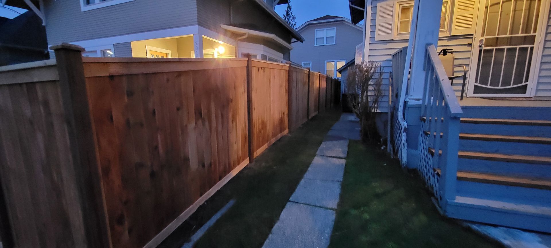 A narrow alleyway between wooden fence and house with blue stairs and a walkway. Evening.