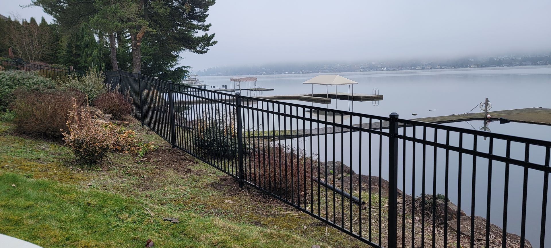 A black metal fence borders a grassy hill overlooking a lake and docks under a cloudy sky.