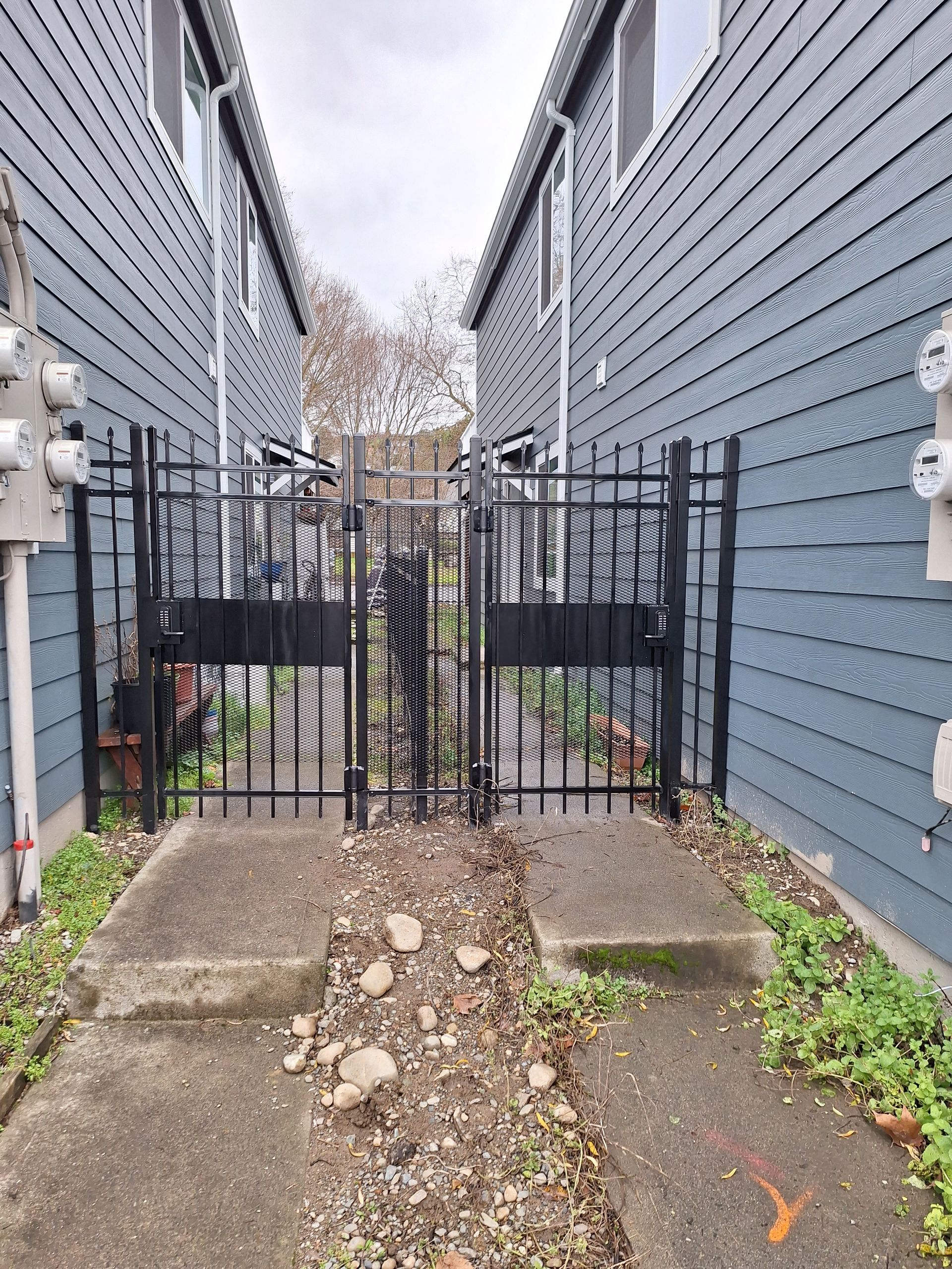 Black iron gate between two blue-sided buildings. Pathway has concrete blocks, gravel, and weeds. Cloudy sky.