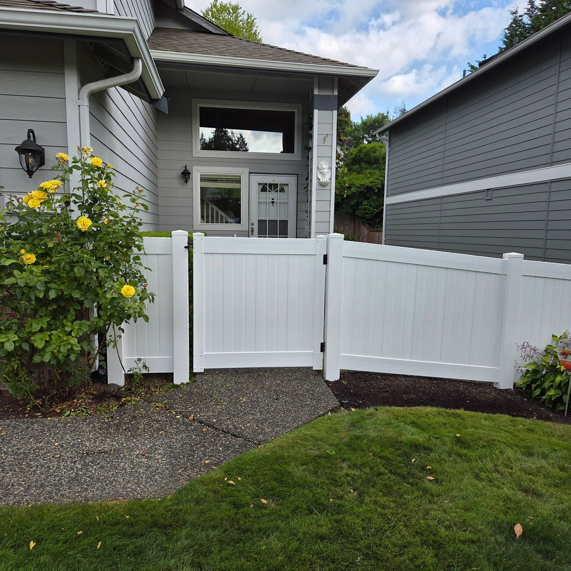 White picket fence in front of a gray house with a small gravel path and grass in the foreground.