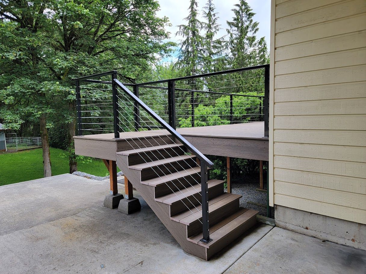 Deck with stairs and cable railing, adjacent to a beige house, with trees in the background.