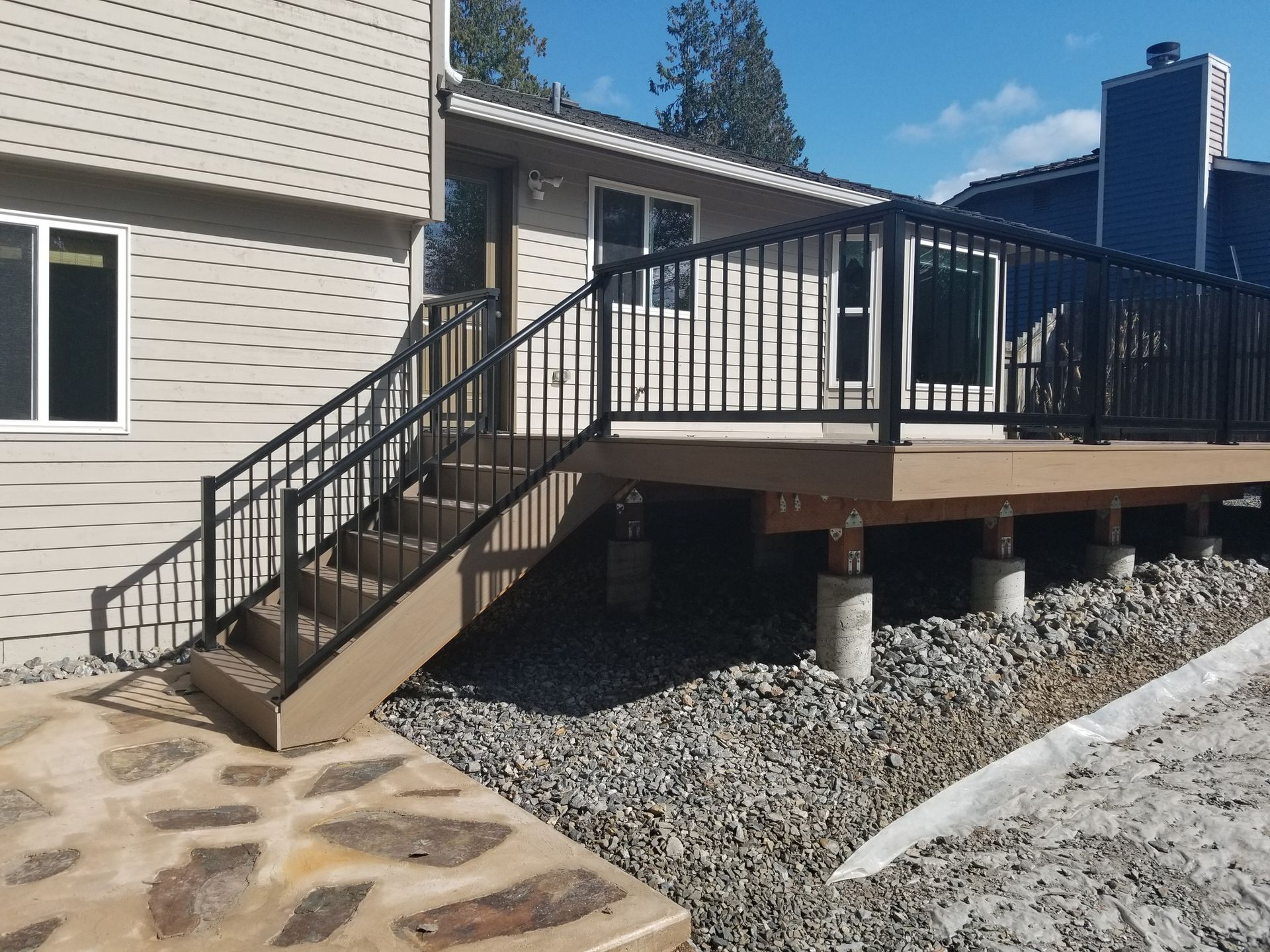 Deck with stairs, black railing, brown composite decking, gravel base, and light beige house.