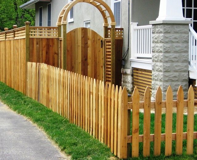 Wooden picket fence along a green lawn and sidewalk, with a gate and arbor leading to a house.