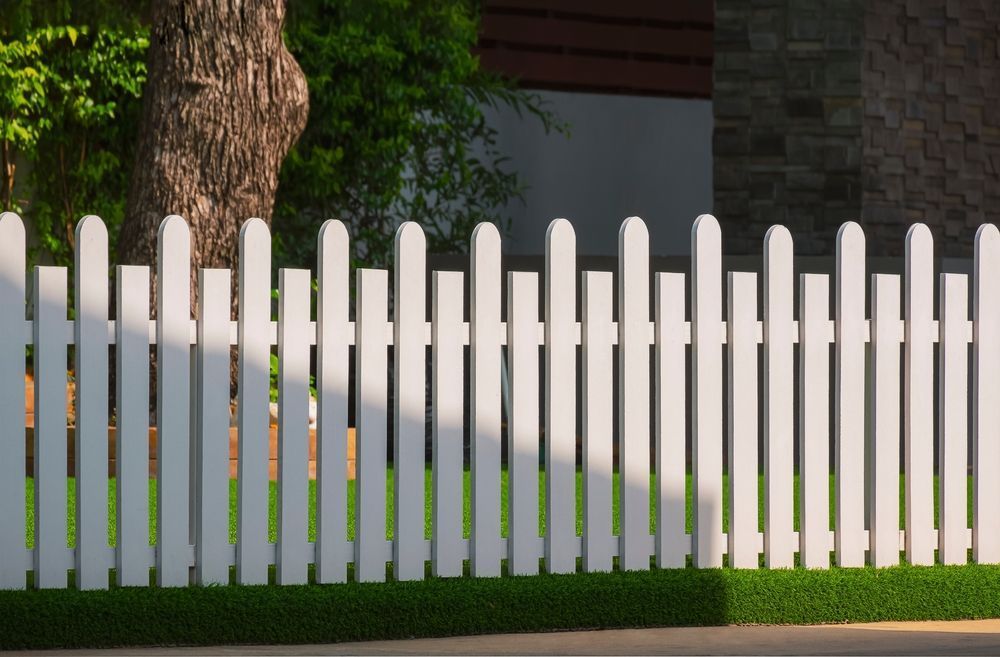 White picket fence in front of green grass, with a tree and part of a house in the background.