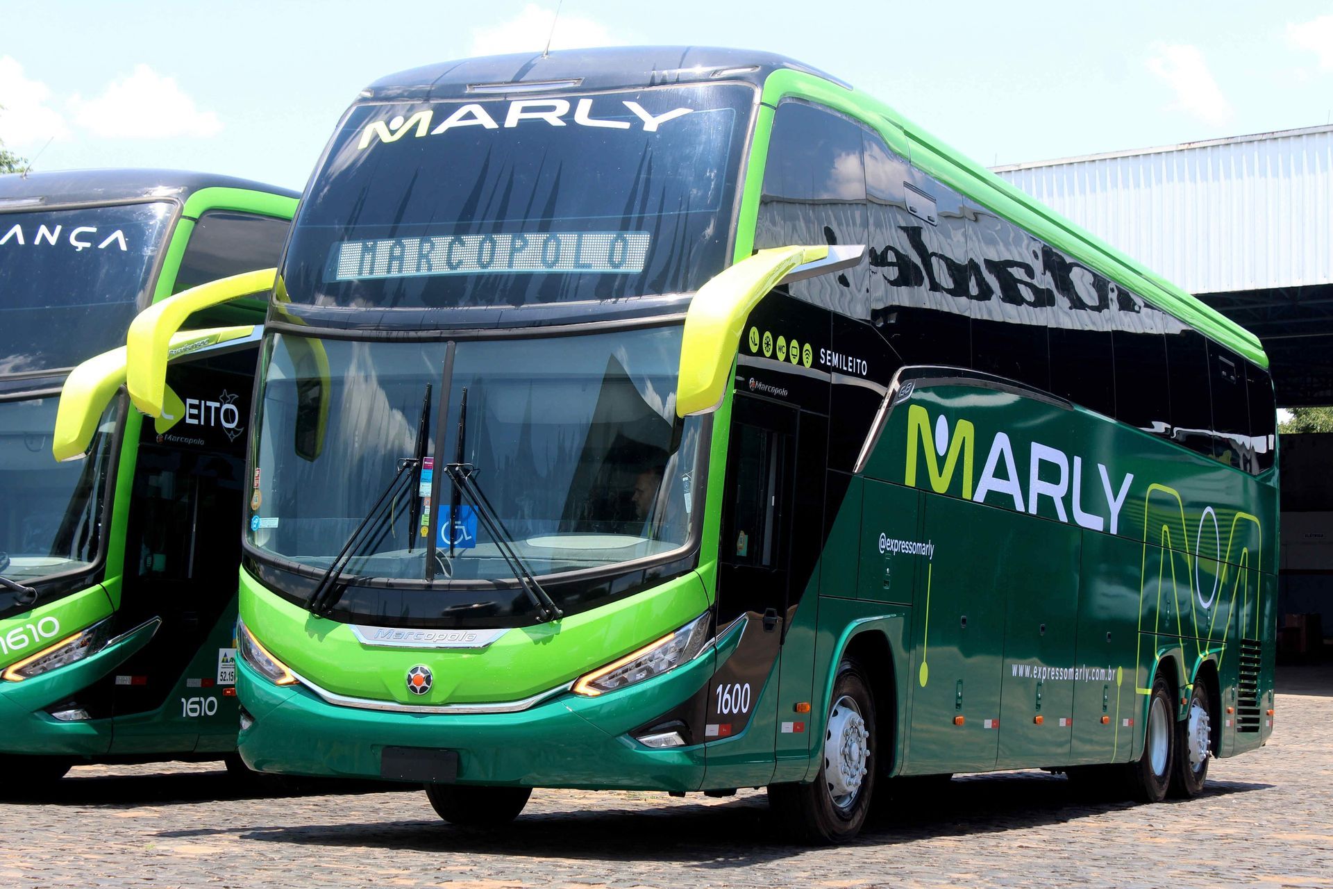Ônibus Marly de dois andares verde e preto estacionado com um ônibus similar, contra um fundo de luz.