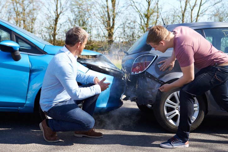 Two men examining damage to cars after a collision on a road. Blue and gray cars, outdoor setting.