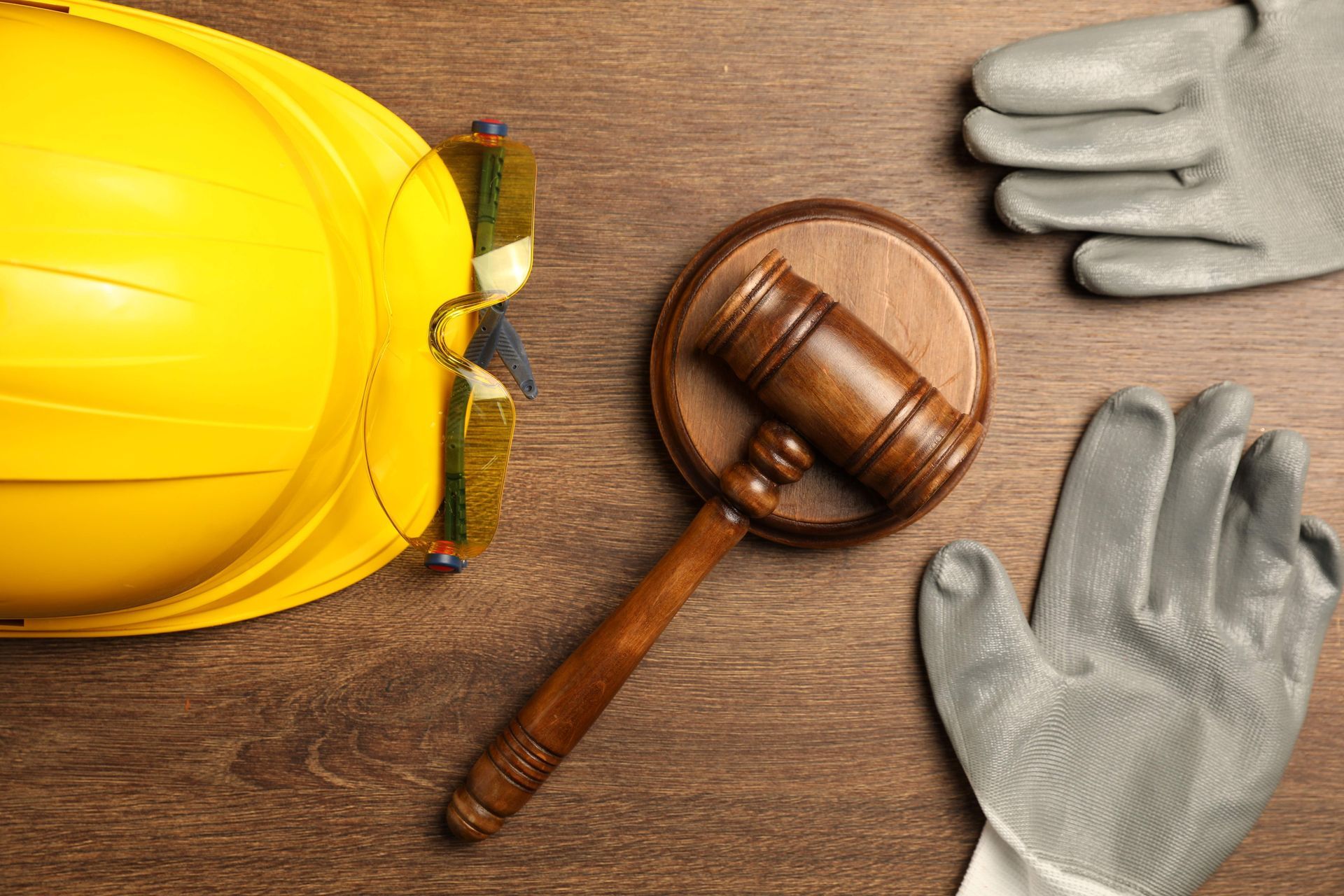 A yellow hard hat, safety gloves, and a wooden gavel displayed together on a wooden surface. A yellow hard hat, safety gloves, and a wooden gavel displayed together on a wooden surface.