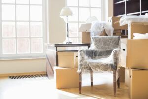 Packed living room with cardboard boxes, a chair wrapped in plastic, and a bright window.