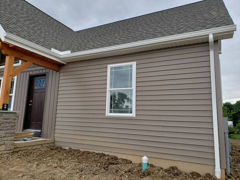 A brown-sided house exterior with white trim, a window, a dark front door, and a white downspout on a dirt lot.