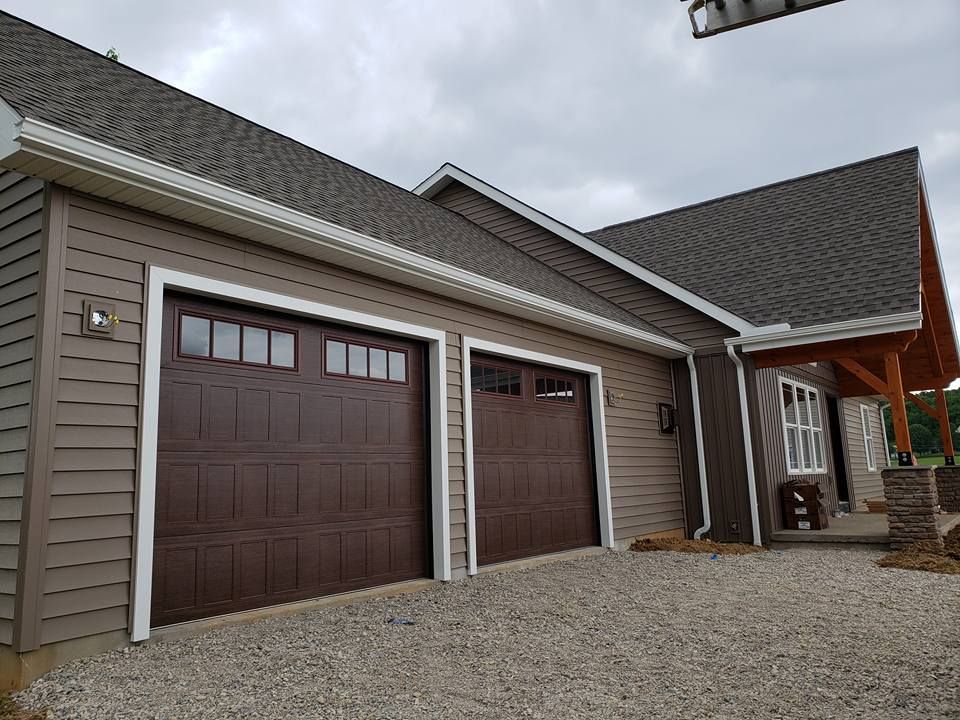 A two-car garage with dark brown doors and windows, attached to a house with beige siding and a dark shingled roof.