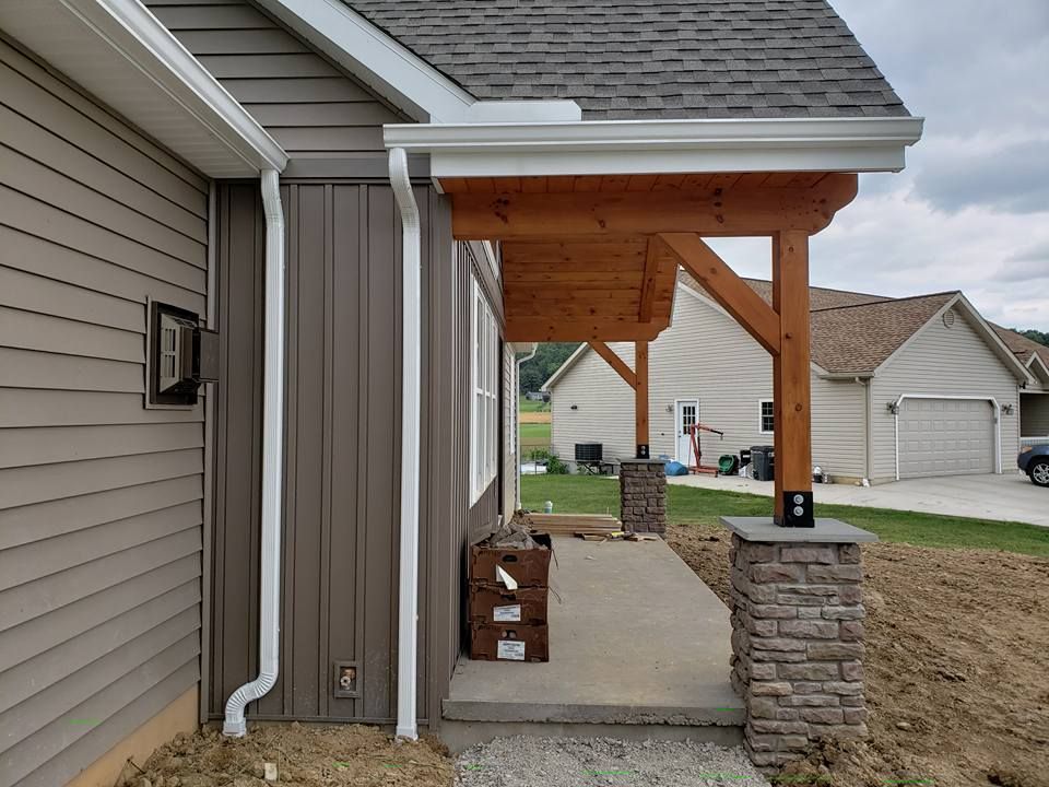 A side view of a house exterior with brown siding, a covered porch with wooden beams, stone pillars, and white gutters.