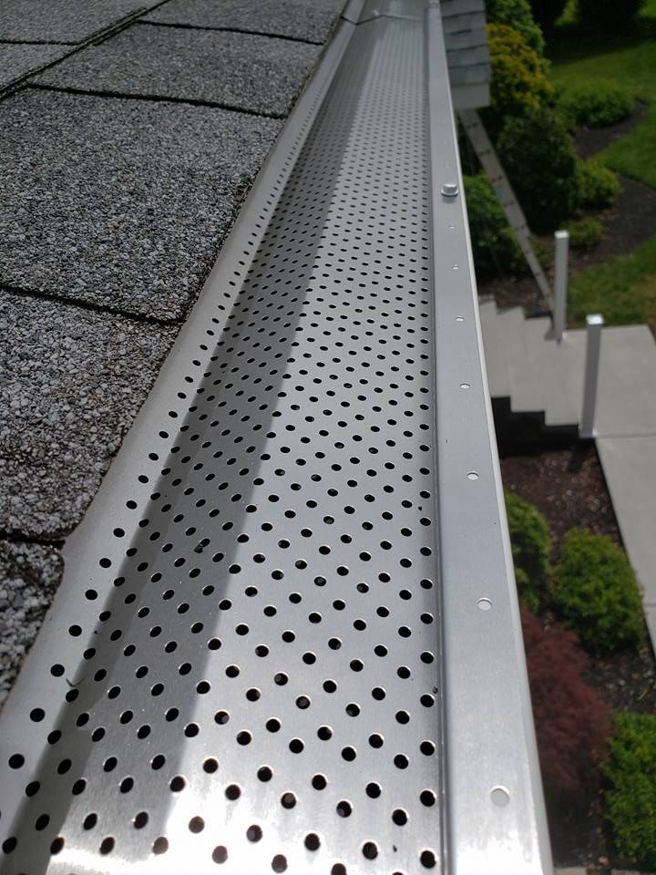 A metal gutter guard with a perforated pattern installed over a roof gutter next to asphalt shingles.