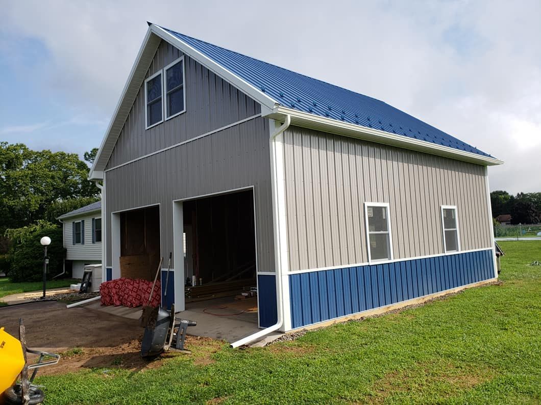 A two-story pole barn with gray vertical siding, a blue metal roof, blue lower trim, and white-framed windows.
