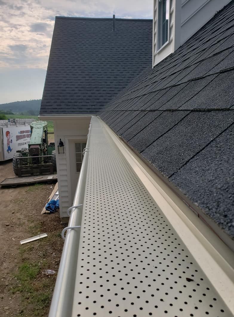 A close-up view of a metal gutter guard installed on a house, with a shingled roof above and construction area nearby.