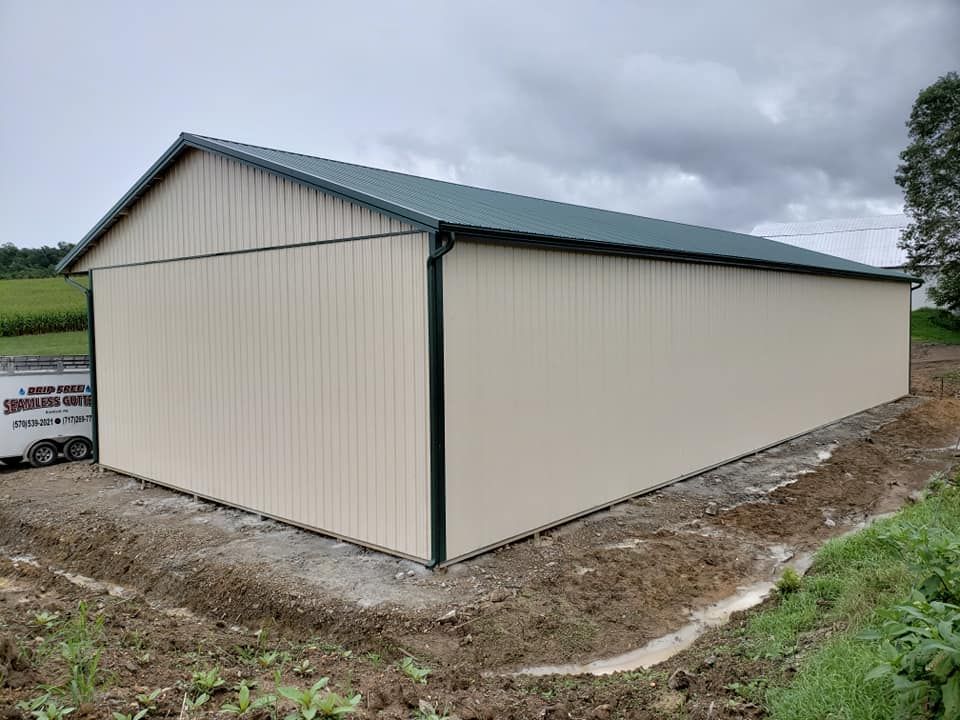 A newly constructed light-beige metal building with a dark green roof, situated on a dirt lot under a cloudy sky.