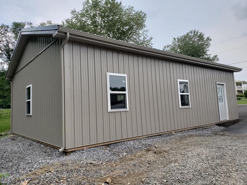 A light tan metal storage building with two windows and a white door, set on a gravel surface under a cloudy sky.
