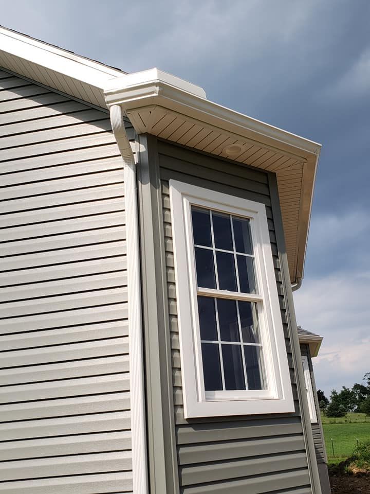 A corner view of a house exterior featuring grey vinyl siding, a white-trimmed window, white soffit, and a white downspout.