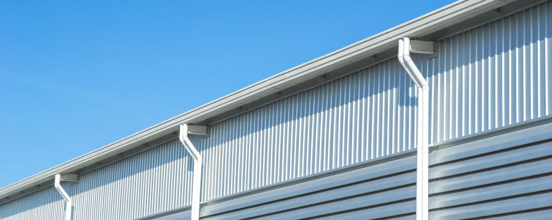 White metal gutters and downspouts attached to a corrugated silver metal industrial building wall under a blue sky.