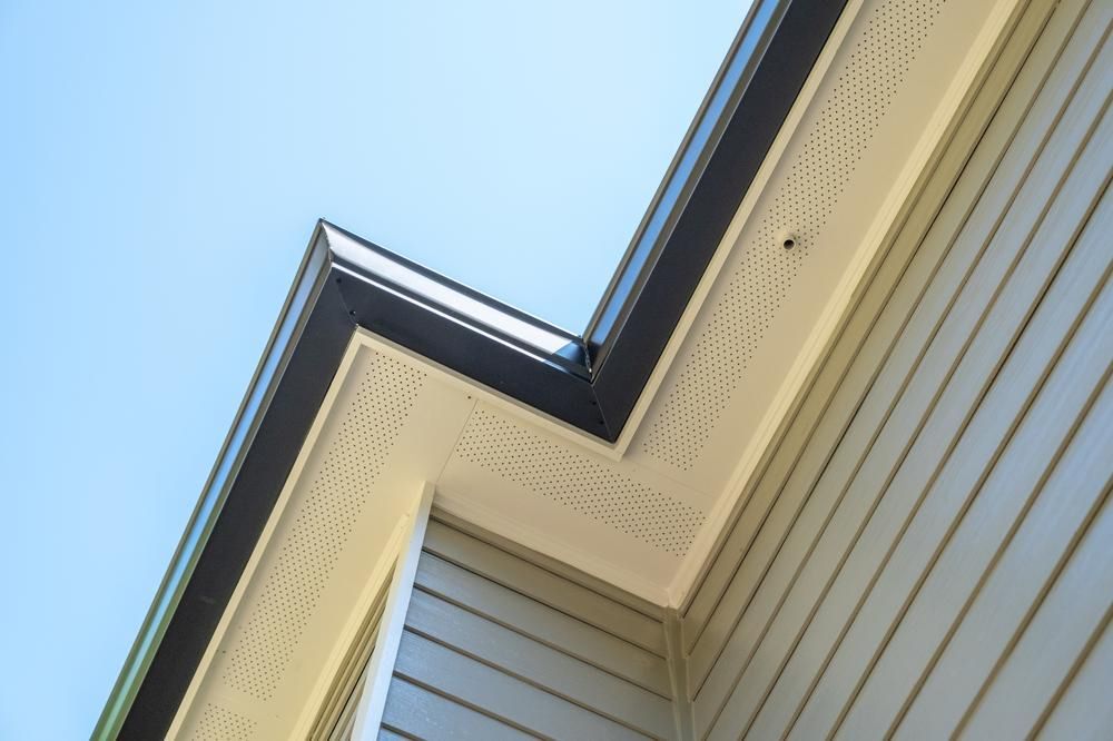 An upward-angled view of a house exterior, showing beige siding, white perforated soffits, & dark fascia against a blue sky.