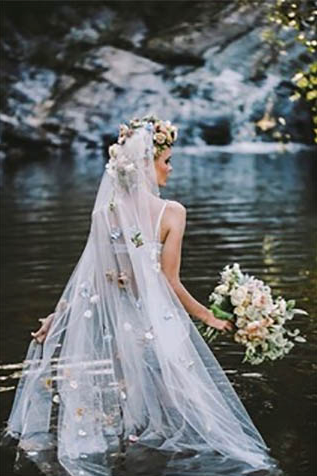 A bride in a wedding dress and veil is standing in the water holding a bouquet of flowers.