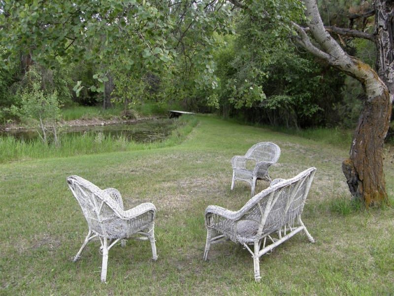 Three white wicker chairs and a bench are sitting in the grass near a pond.