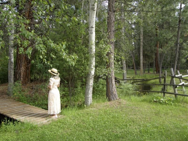 A woman in a white dress and hat is standing on a wooden bridge in the woods.