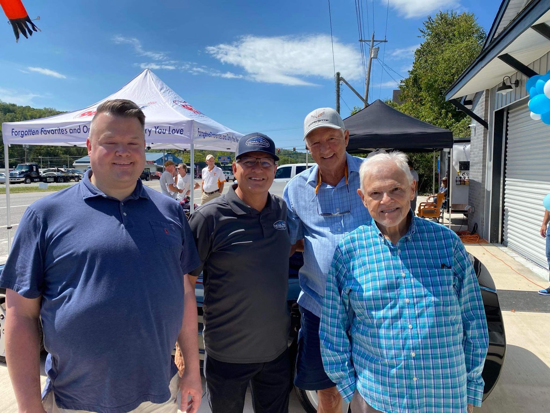 Four men pose outdoors near a tent and building. One wears a blue shirt, others wear caps and casual attire.