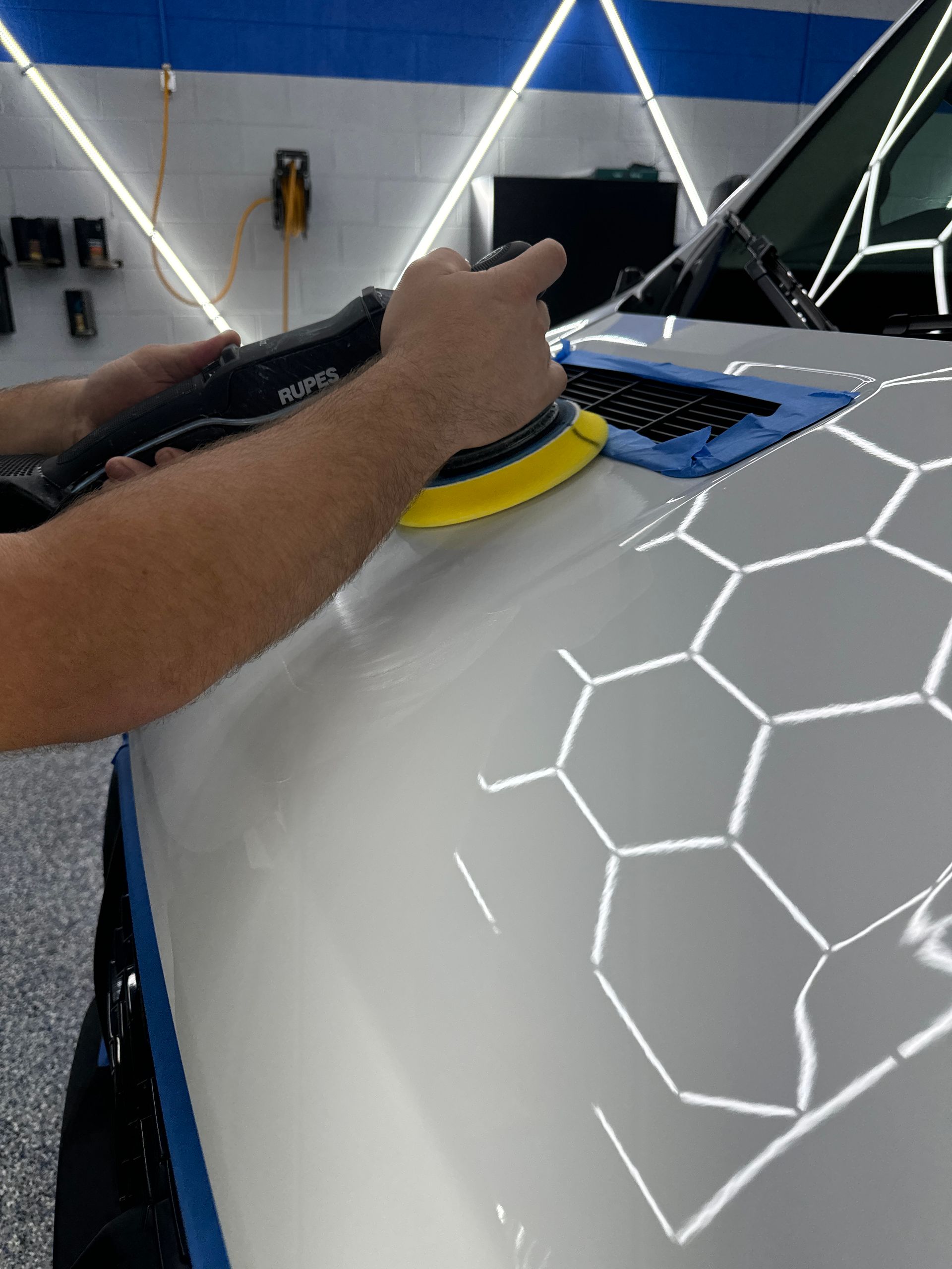 Person polishing white car hood with a machine, reflecting bright lights.