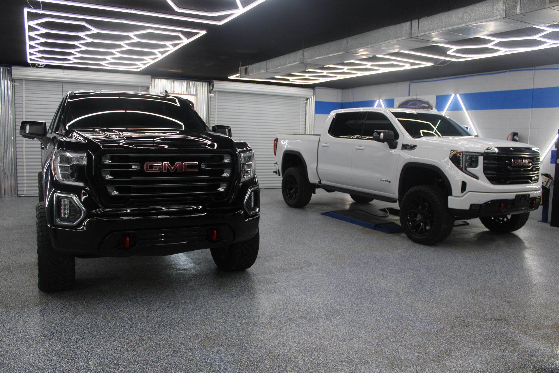 Two GMC Sierra trucks in a garage; one black, one white. Both are lifted with black wheels.