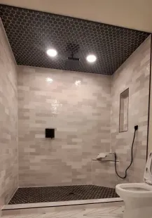 Shower stall with gray and black tiled walls and ceiling. Black stone floor. White toilet in foreground.