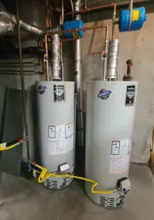 Two gas water heaters in a basement, connected by copper pipes. Yellow gas lines and blue expansion tanks are visible.