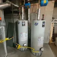 Two connected water heaters in a basement. Copper pipes and blue expansion tanks are visible.