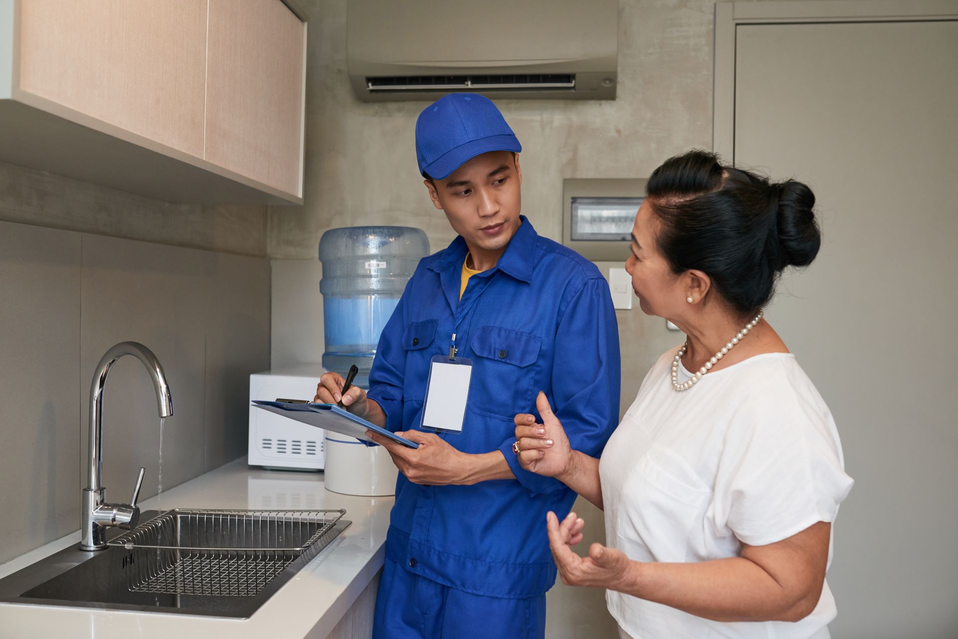 Plumber in blue uniform, taking notes while talking to woman in a kitchen with sink and water cooler.