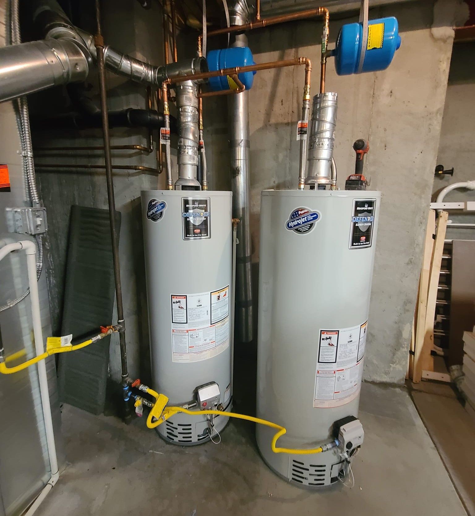 Two connected water heaters in a basement. Copper pipes and blue expansion tanks are visible.