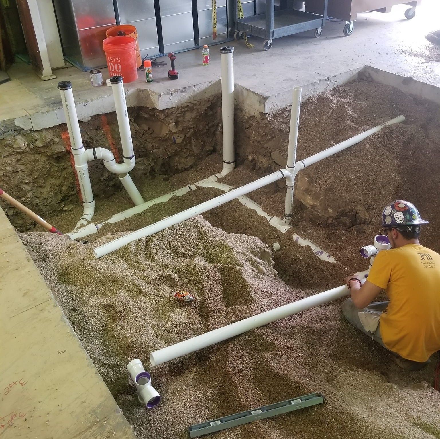 Plumber installing PVC pipes in a gravel-filled trench. A worker sits nearby, holding a pipe.