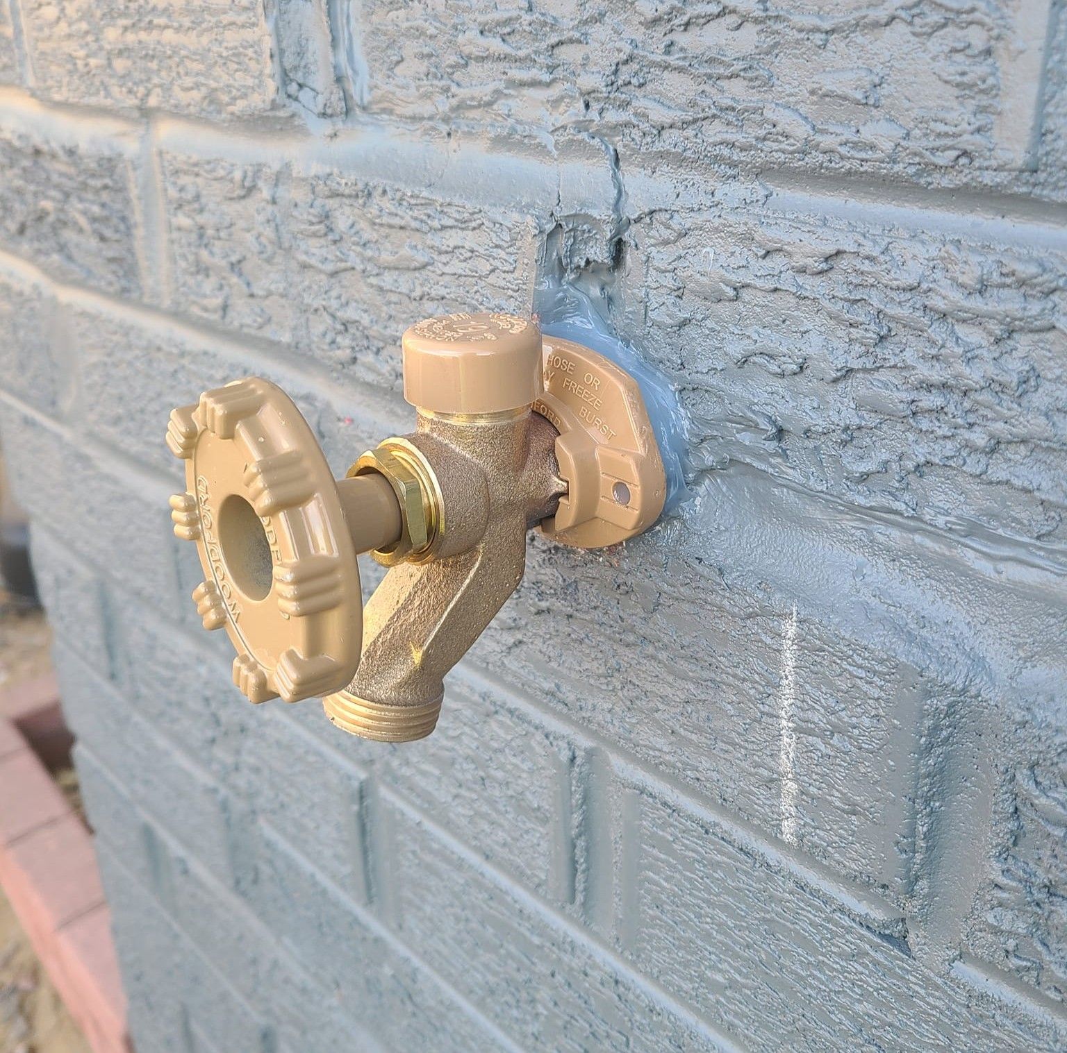 Tan outdoor water faucet mounted on a painted gray brick wall.