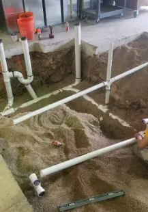Plumber installs white PVC pipes in a trench filled with gravel. He wears a yellow shirt and is working near a concrete slab.