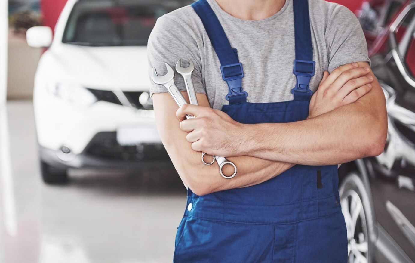 A man in blue overalls is holding a wrench in front of a car.