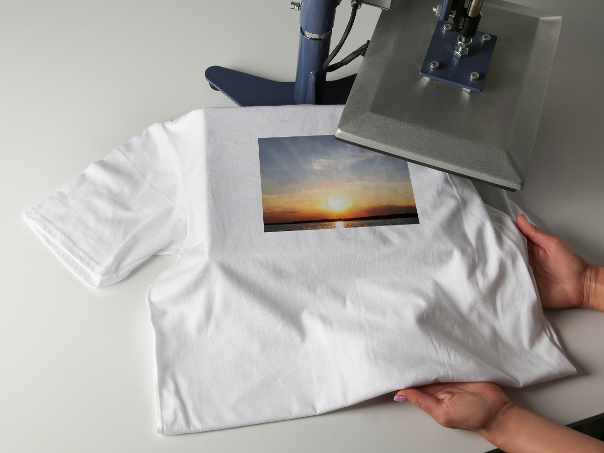 White t-shirt with a sunset design being pressed by a heat press. Hands hold the shirt on a white surface. White t-shirt with a sunset design being pressed by a heat press. Hands hold the shirt on a white surface.